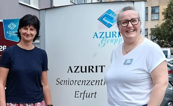 Zwei lächelnde Frauen stehen vor einem Schild mit der Aufschrift 'AZURIT Seniorenzentrum Erfurt' und einem blauen Logo