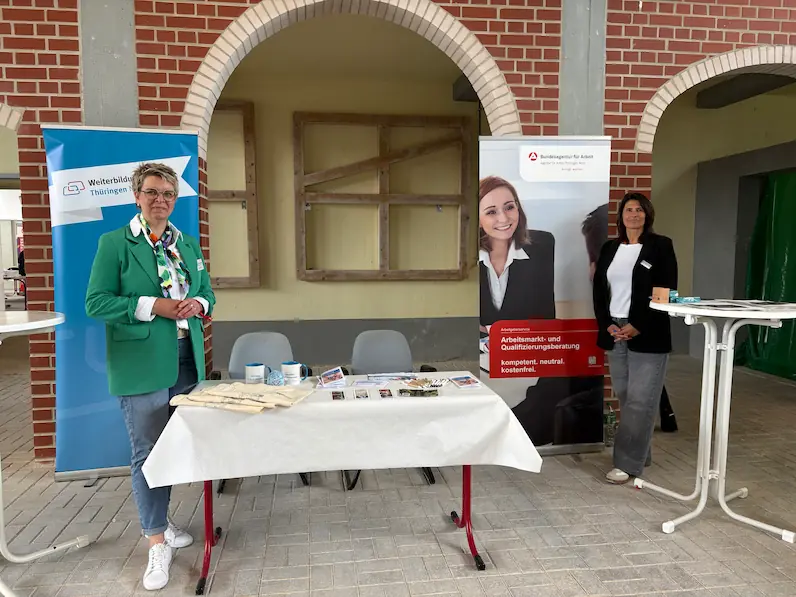 Zwei Frauen stehen an einem mit Informationsmaterial gedeckten Tisch vor einem Backsteinbogen, links ein Banner der Weiterbildungsagentur Thüringen, rechts ein Banner der Bundesagentur für Arbeit.