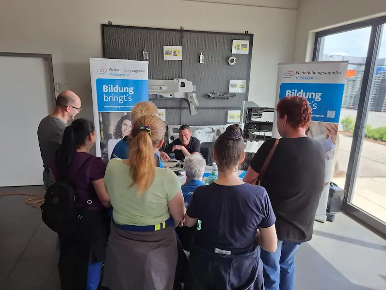 Gruppe von Menschen steht vor einem Tisch mit zwei Bannern der Weiterbildungsagentur Thüringen Ost und einem Mann, der am Tisch sitzt und spricht.