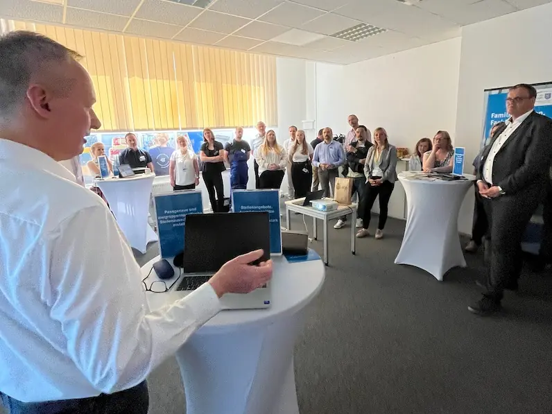 Mann im weißen Hemd hält Vortrag vor sitzender und stehender Gruppe in hellem Konferenzraum mit Stehtischen
