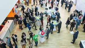 Menschenmenge auf einer Messe zur Weiterbildung, verschiedene Stände mit Tischen in unterschiedlichen Farben, Teilnehmer in formeller Kleidung.