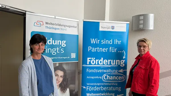 Anke Lettau und Katja Kühn stehen vor Informationsaufstellern der Weiterbildungsagentur Thüringen und des Thüringer Landesverwaltungsamts.