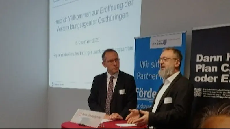 Zwei Männer in Anzügen stehen an einem hohen, rot bezogenen Stehtisch vor einer Wand mit einer Projektionsfläche und einem Banner der Weiterbildungsagentur Thüringen Ost