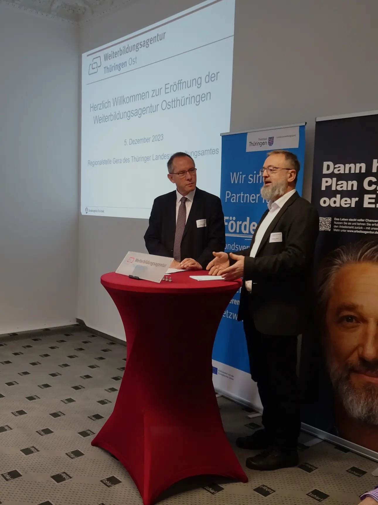 Zwei Männer in Anzügen stehen an einem hohen, rot bezogenen Stehtisch vor einer Wand mit einer Projektionsfläche und einem Banner der Weiterbildungsagentur Thüringen Ost