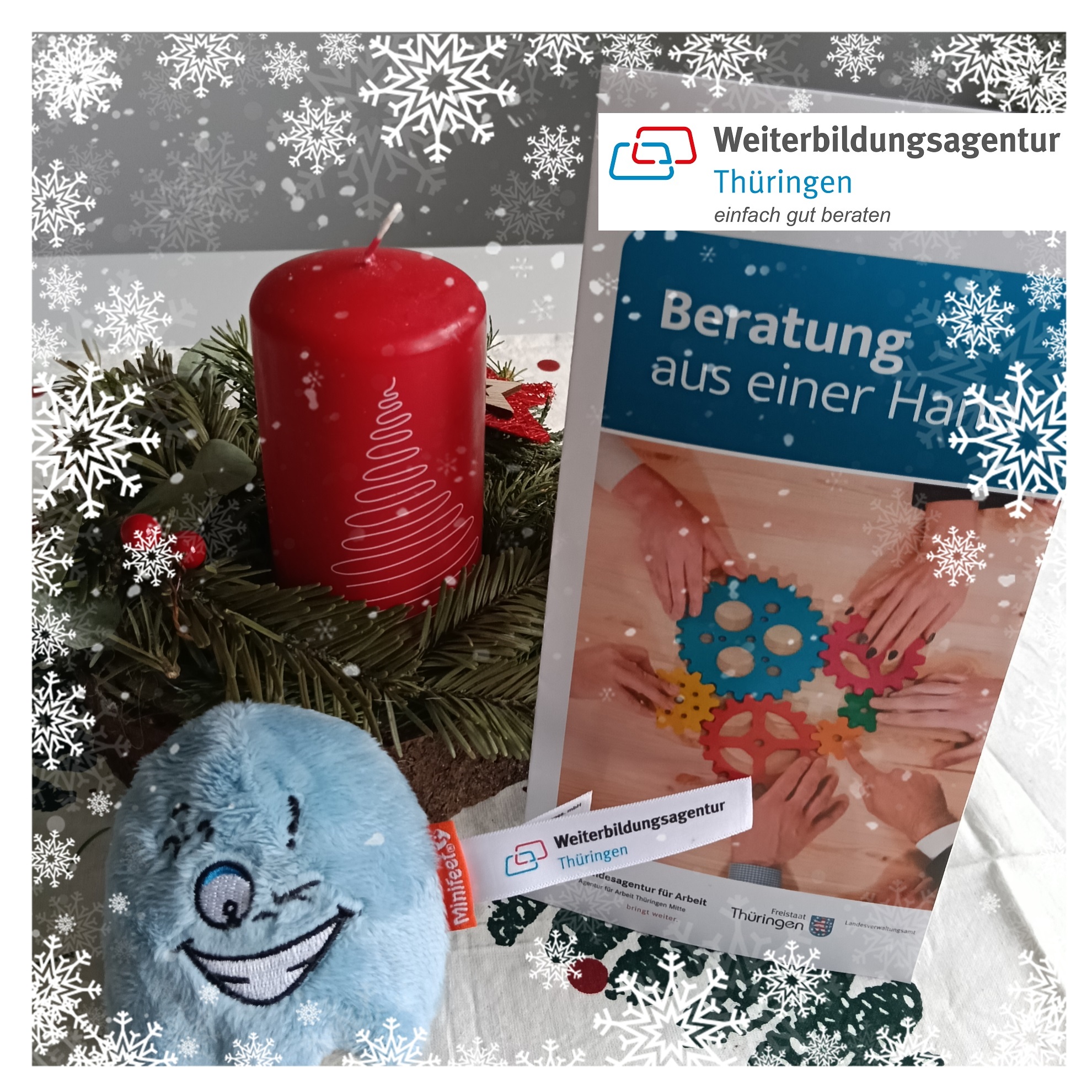 Kerzenständer mit Tannenzweigen, roter Kerze und einem blauen, lachenden Plüschgesicht neben einem Flyer der Weiterbildungsagentur Thüringen mit bunten Zahnrädern und dem Text 'Beratung aus einer Hand'.