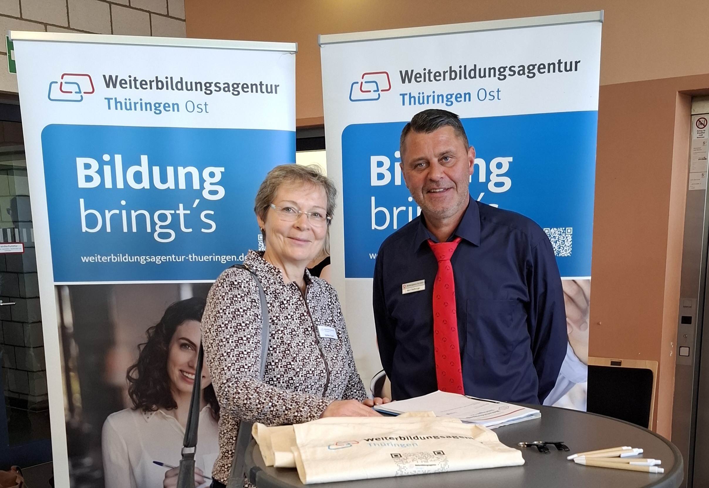 Zwei Personen stehen an einem runden Stehtisch. Im Hintergrund sind zwei Banner der Weiterbildungsagentur Thüringen Ost mit dem Text 'Bildung bringt's' zu sehen.
