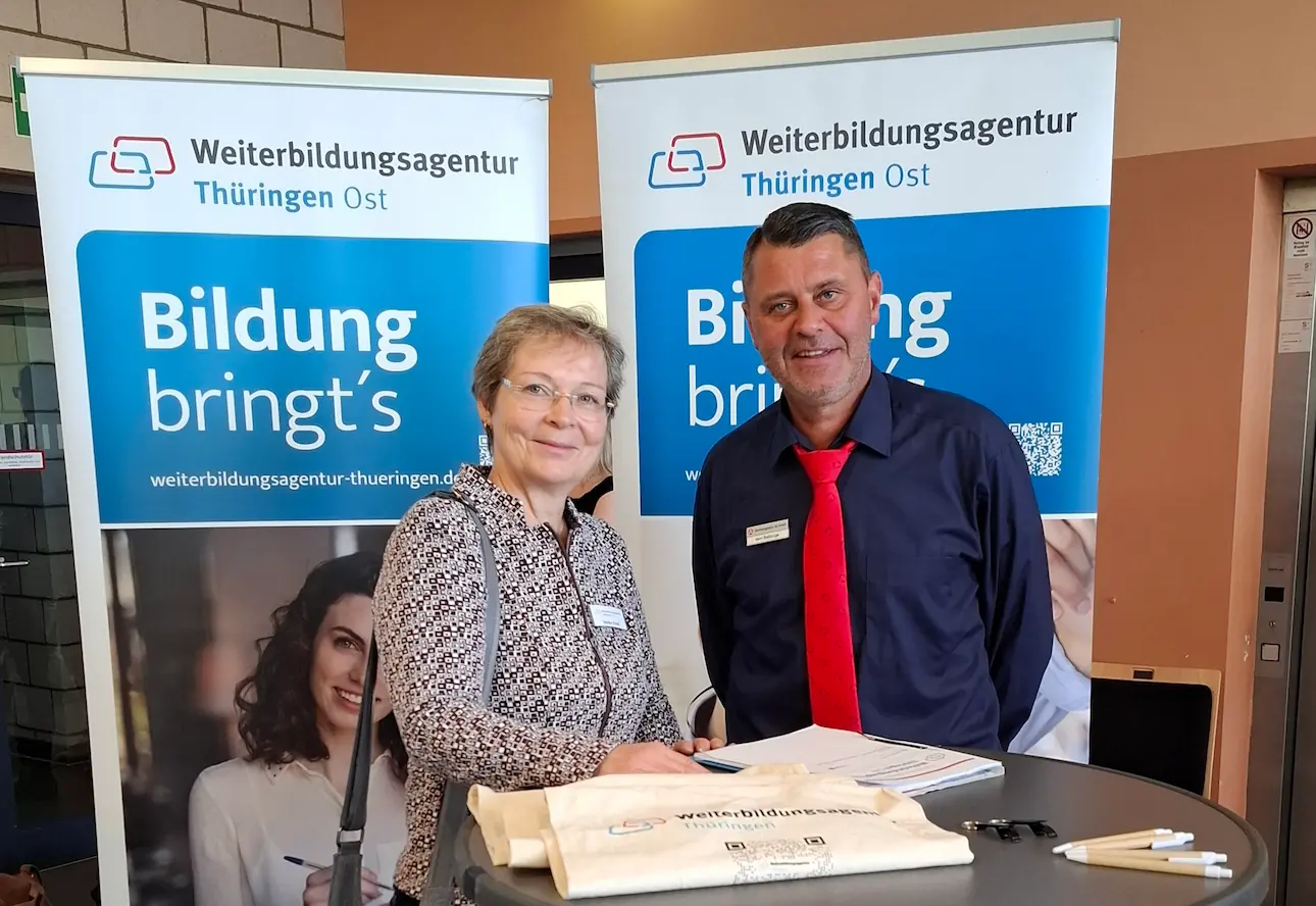Zwei Personen stehen an einem runden Stehtisch. Im Hintergrund sind zwei Banner der Weiterbildungsagentur Thüringen Ost mit dem Text 'Bildung bringt's' zu sehen.