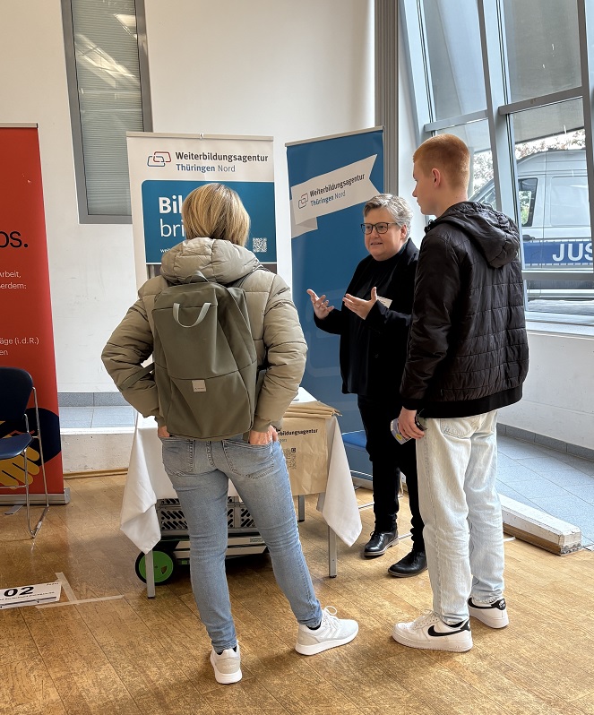 Zwei junge Personen mit Jacken und Rucksäcken stehen vor einem Stand der Weiterbildungsagentur Thüringen Nord, eine Person erklärt etwas mit Gestik.