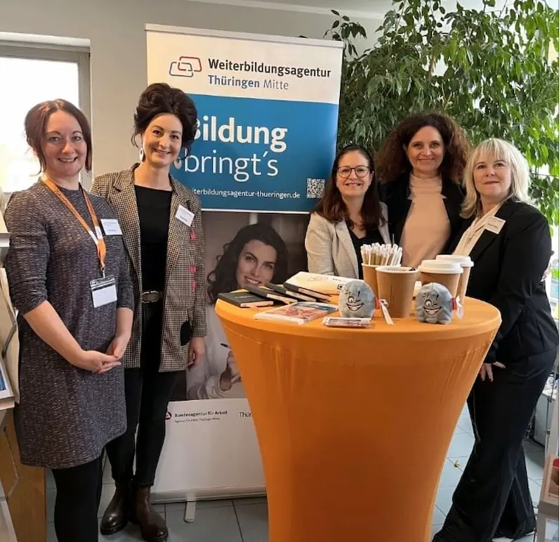 Fünf Beraterinnen stehen an einem orangefarbenen Stehtisch mit Werbematerial. Im Hintergrund ein Banner der Weiterbildungsagentur Thüringen  mit dem Text "Bildung bringt's".