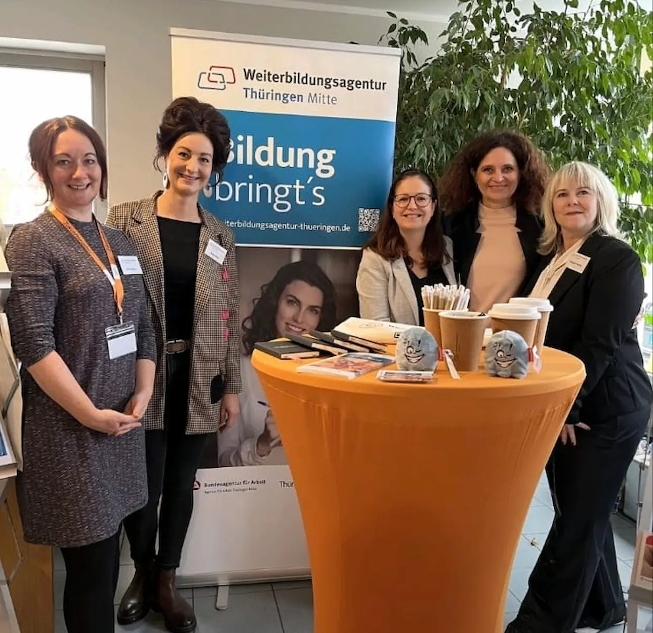 Fünf Beraterinnen stehen an einem orangefarbenen Stehtisch mit Werbematerial. Im Hintergrund ein Banner der Weiterbildungsagentur Thüringen  mit dem Text "Bildung bringt's".