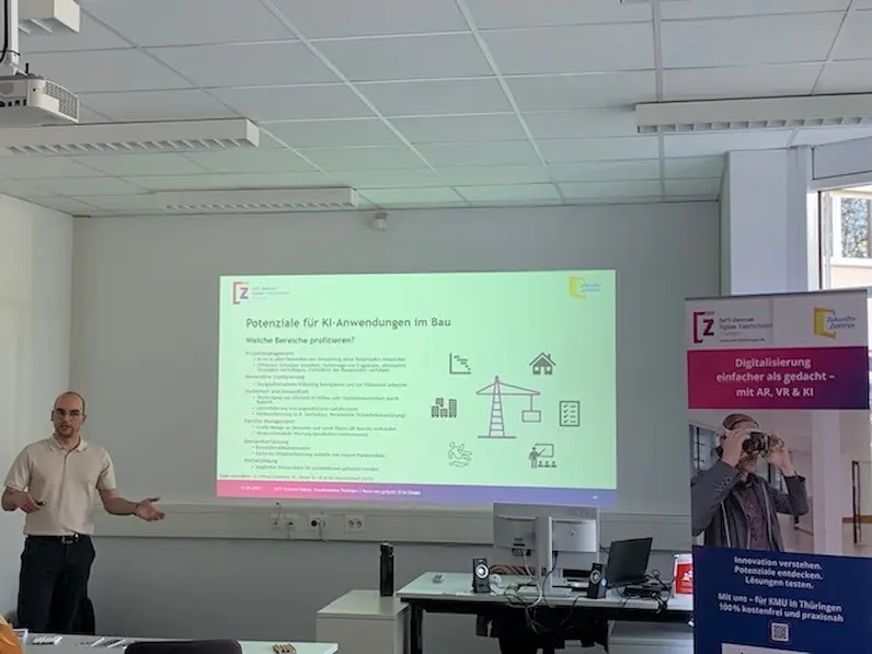Vortragender steht links vor einer Präsentationsfolie mit dem Titel 'Potenziale für KI-Anwendungen im Bau' in einem Seminarraum, rechts steht ein Roll-up des ZeTT mit dem Titel "Digitalisierung einfacher als gedacht - mit AR, VR und KI"