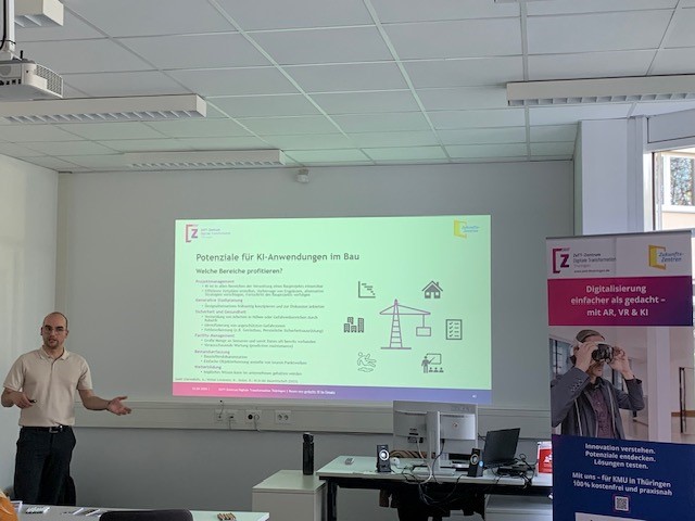Vortragender steht links vor einer Präsentationsfolie mit dem Titel 'Potenziale für KI-Anwendungen im Bau' in einem Seminarraum, rechts steht ein Roll-up des ZeTT mit dem Titel 