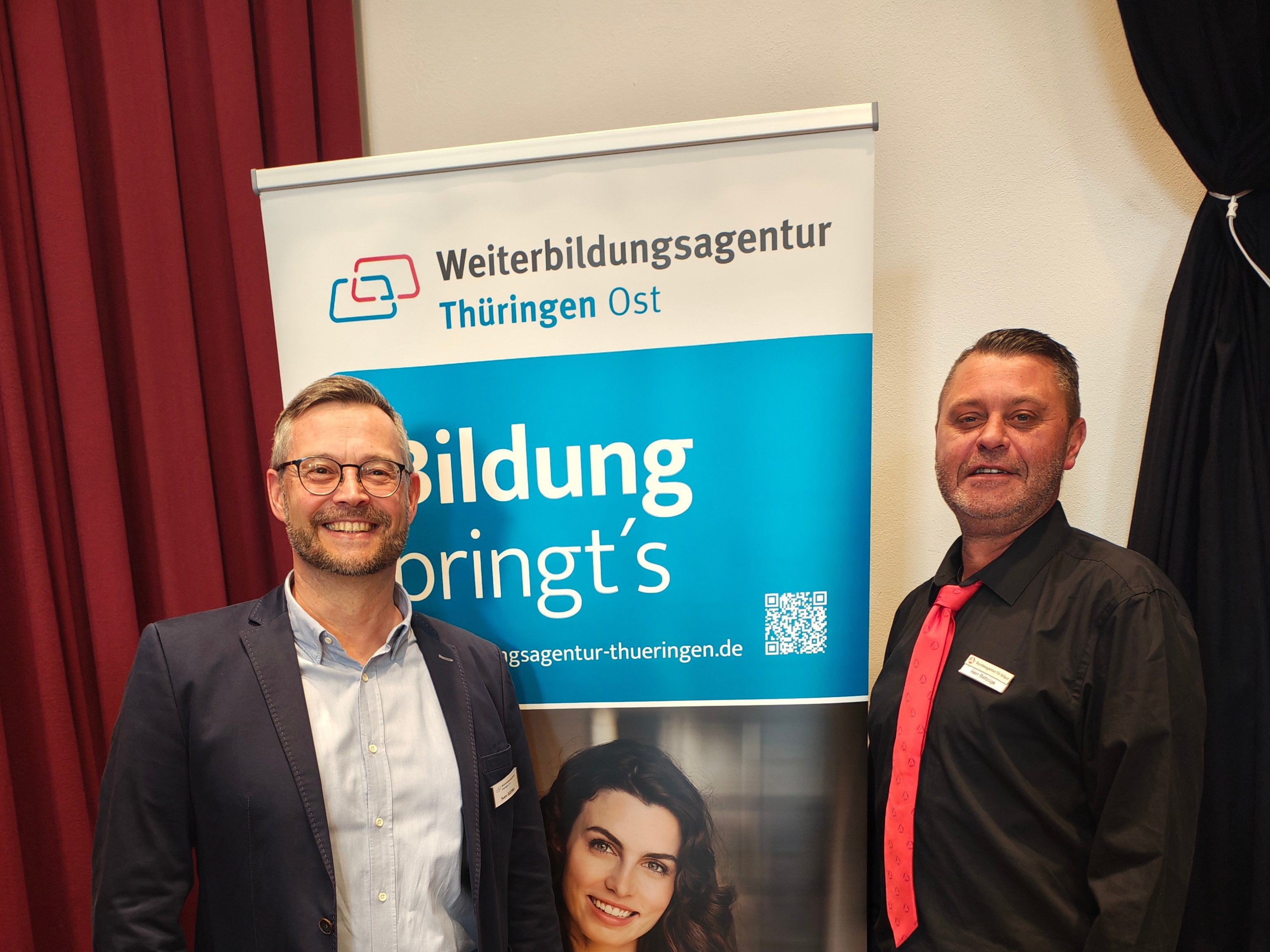 Zwei Männer stehen vor einem Roll-up mit der Aufschrift 'Weiterbildungsagentur Thüringen Ost Bildung bringt's' in einem Raum mit Holzboden und rotem Vorhang