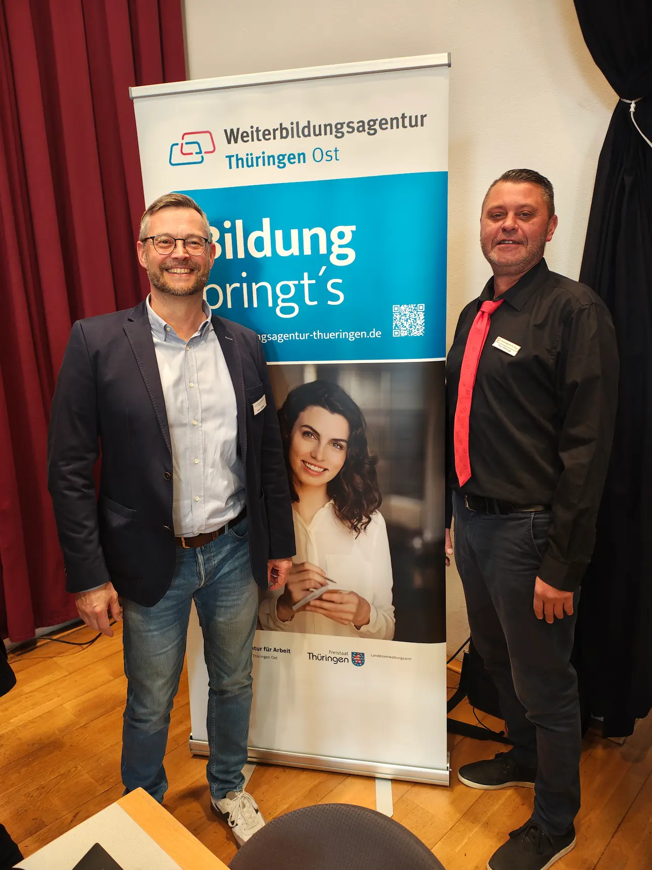 Zwei Männer stehen vor einem Roll-up mit der Aufschrift 'Weiterbildungsagentur Thüringen Ost Bildung bringt's' in einem Raum mit Holzboden und rotem Vorhang