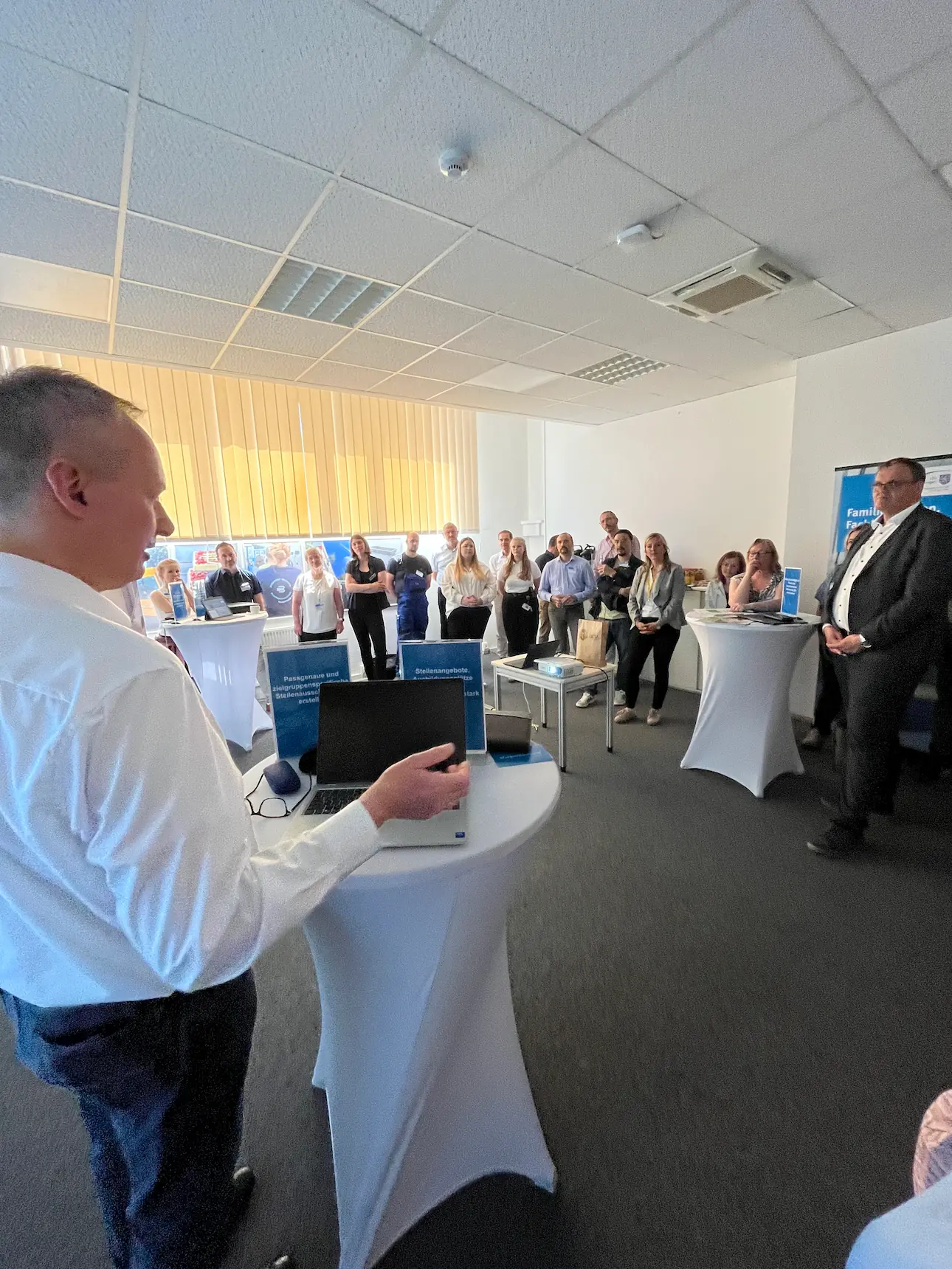 Mann im weißen Hemd hält Vortrag vor sitzender und stehender Gruppe in hellem Konferenzraum mit Stehtischen