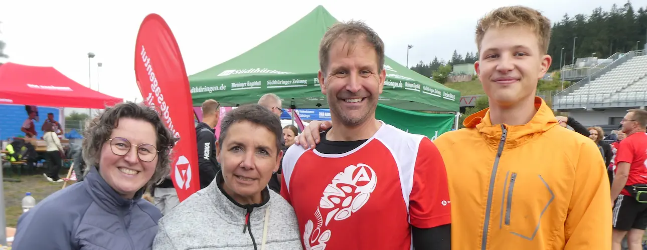 Vier Personen posieren beim 16. Firmenlauf Thüringens Süden in Oberhof vor Veranstaltungszelten und einer Tribüne; ein Teilnehmer trägt ein rot-weißes Laufshirt.