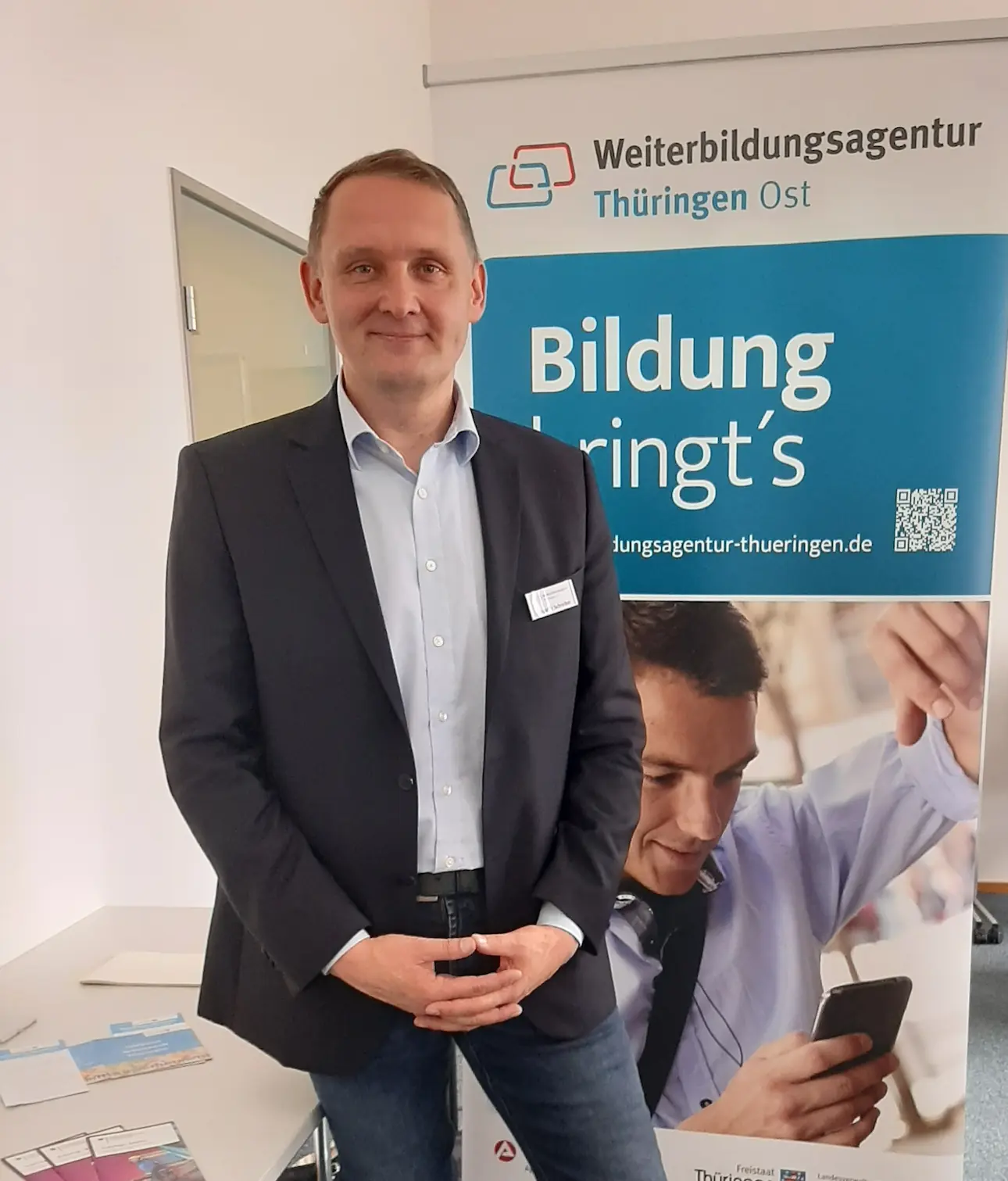 Ein Mann in dunklem Sakko und Jeans steht vor einem Roll-up mit der Aufschrift "Weiterbildungsagentur Thüringen Ost Bildung bringt's" in einem Büro mit Stuhl und Tisch im Hintergrund