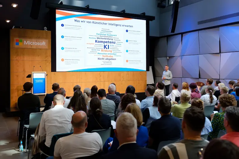 Vortragender steht vor Publikum in modernem Konferenzraum und präsentiert Folie zum Thema Künstliche Intelligenz