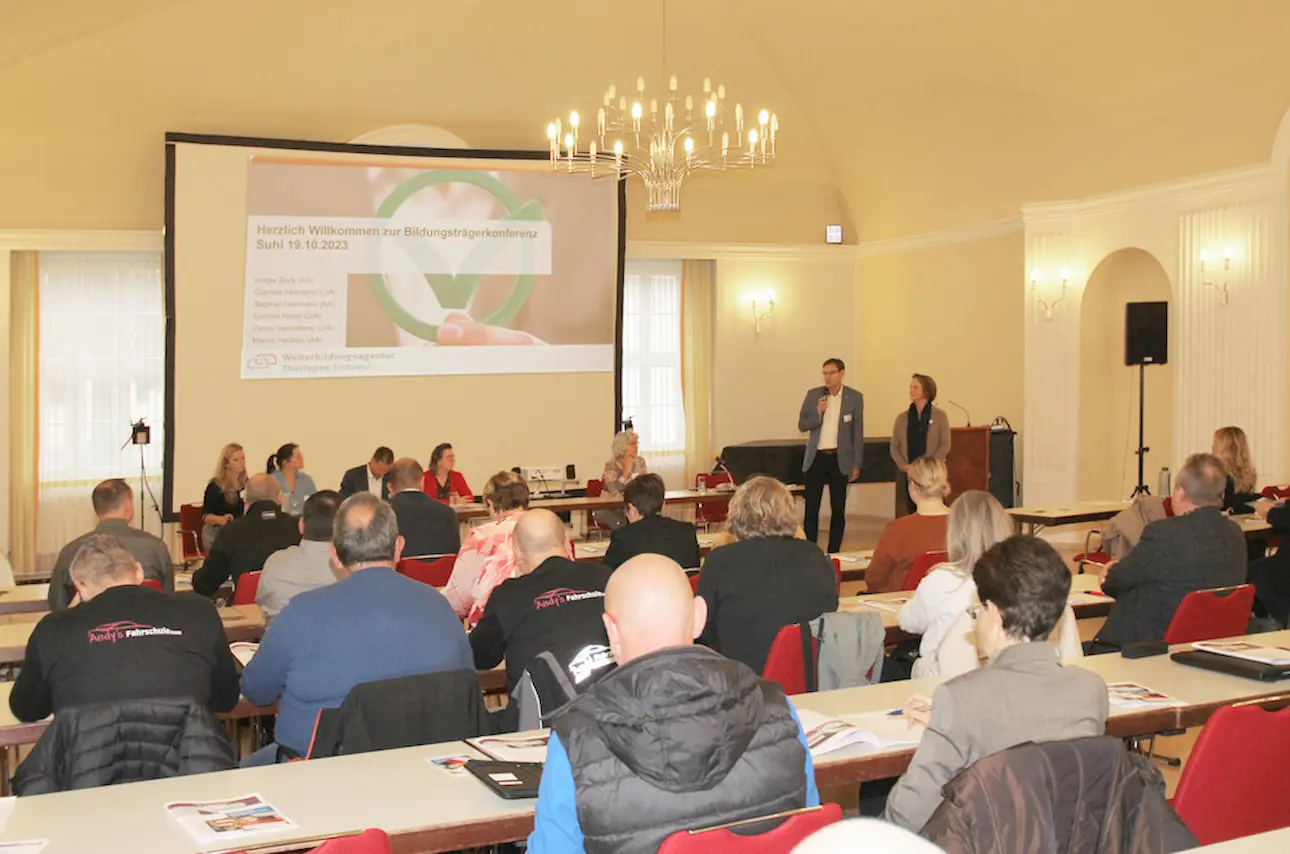 Mehrere Personen sitzen in einem Saal mit Kronleuchtern und hören zwei stehenden Vortragenden vor einer Leinwand mit Präsentation zu