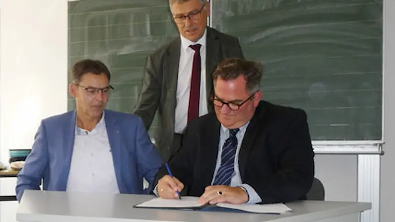 Drei Männer in Anzügen sitzen und stehen an einem weißen Tisch vor einer grünen Tafel, einer unterschreibt ein Dokument.