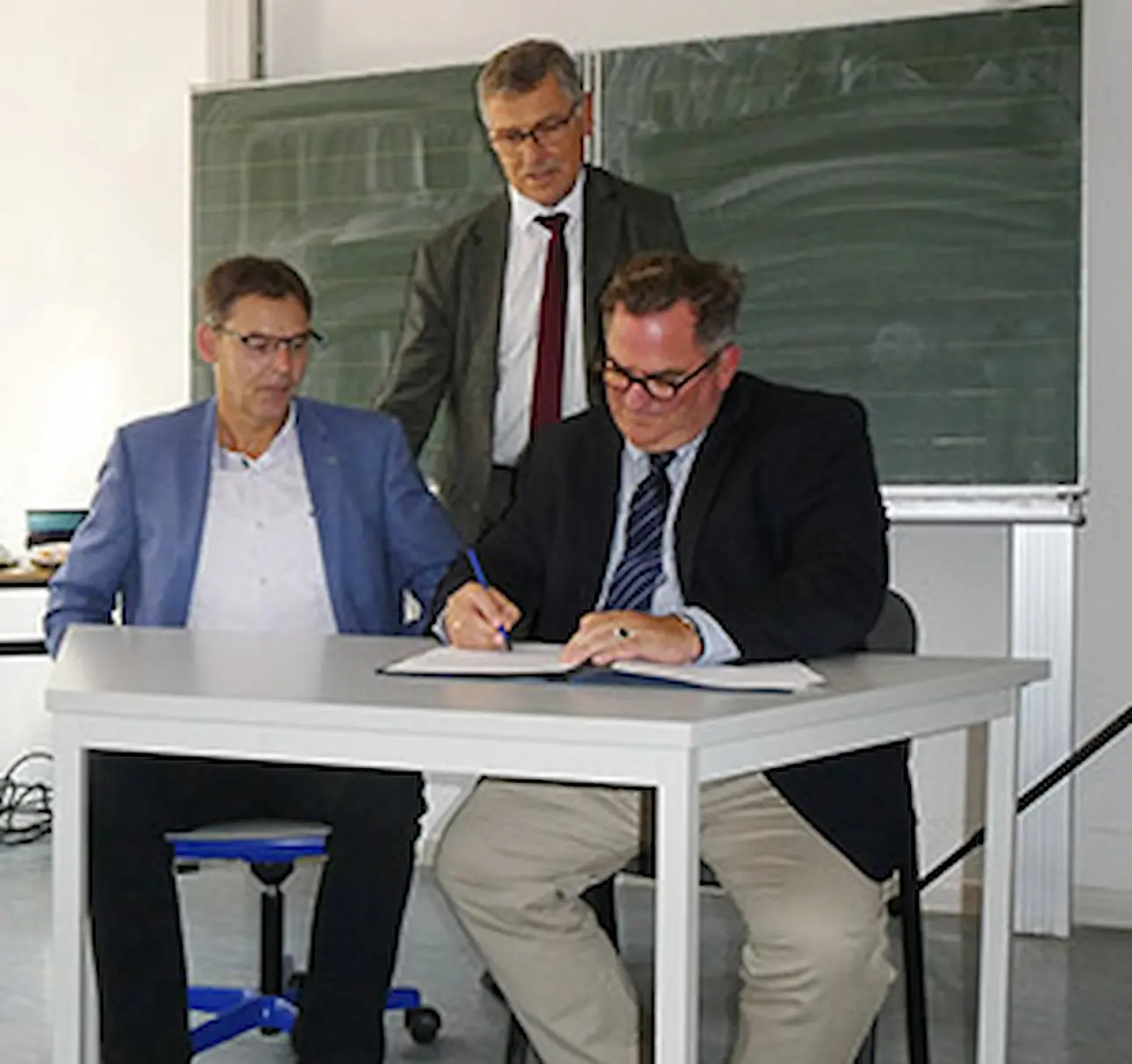 Drei Männer in Anzügen sitzen und stehen an einem weißen Tisch vor einer grünen Tafel, einer unterschreibt ein Dokument.