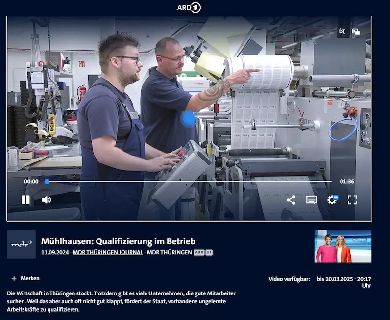 Zwei Männer in Arbeitskleidung bedienen eine Maschine mit Papierrolle in einer Produktionshalle