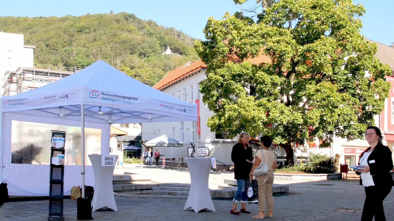 Menschen an Stehtischen unter einem Pavillon mit Aufschrift zur Weiterbildungsagentur Thüringen Südwest in der Suhler Innenhof mit Bäumen und Gebäuden im Hintergrund