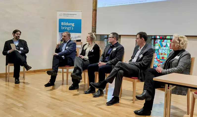 Fünf Personen sitzen in einem Halbkreis auf Stühlen in einem Raum mit Holzboden und diskutieren, im Hintergrund ein Banner mit der Aufschrift 'Weiterbildungsagentur Thüringen Südwest Bildung bringt's'