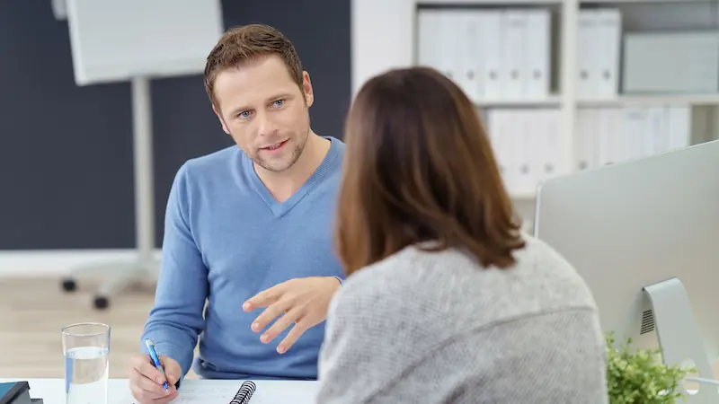 Zwei Personen sitzen sich in einem Büro gegenüber, eine Person mit blauem Pullover erklärt etwas zu Weiterbildung, die andere Person mit braunen Haaren hört zu, auf dem Tisch Notizbuch und Glas Wasser.