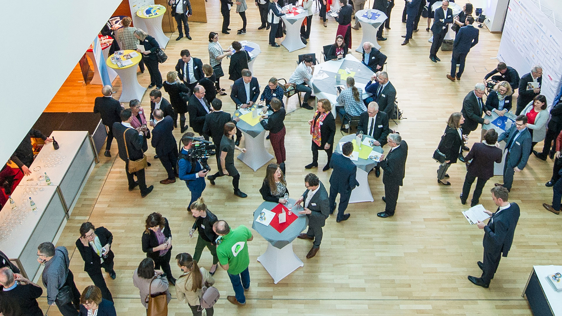 Menschenmenge auf einer Messe zur Weiterbildung, verschiedene Stände mit Tischen in unterschiedlichen Farben, Teilnehmer in formeller Kleidung.